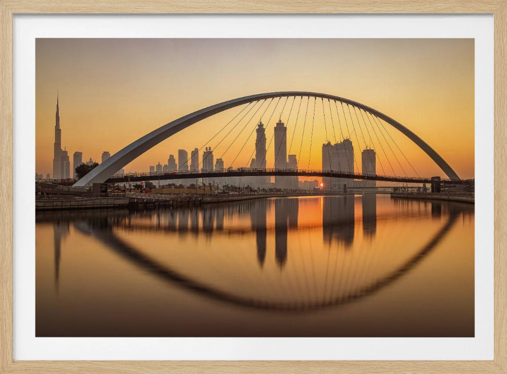 A panoramic photograph of the Dubai skyline at sunrise, viewed from across the water. A large, modern arched bridge spans the canal in the foreground, and its reflection creates a perfect oval shape in the calm water. In the background, skyscrapers, including the Burj Khalifa, are silhouetted against a glowing orange and yellow sky. The entire scene is presented within a silver picture frame. Wall Art