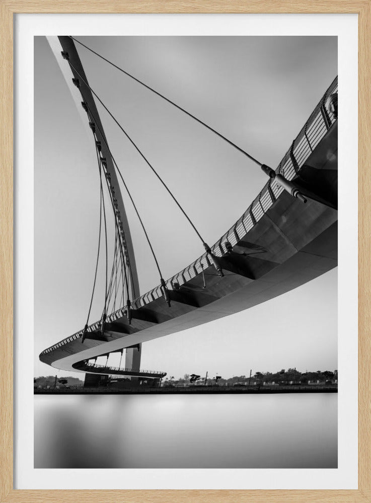 A dynamic low-angle, black and white photograph of a modern cable-stayed pedestrian bridge. The bridge curves elegantly over calm, reflective water, with its deck suspended by numerous cables attached to a tall, angled pylon, creating a striking graphic composition against a bright, clear sky. Decor