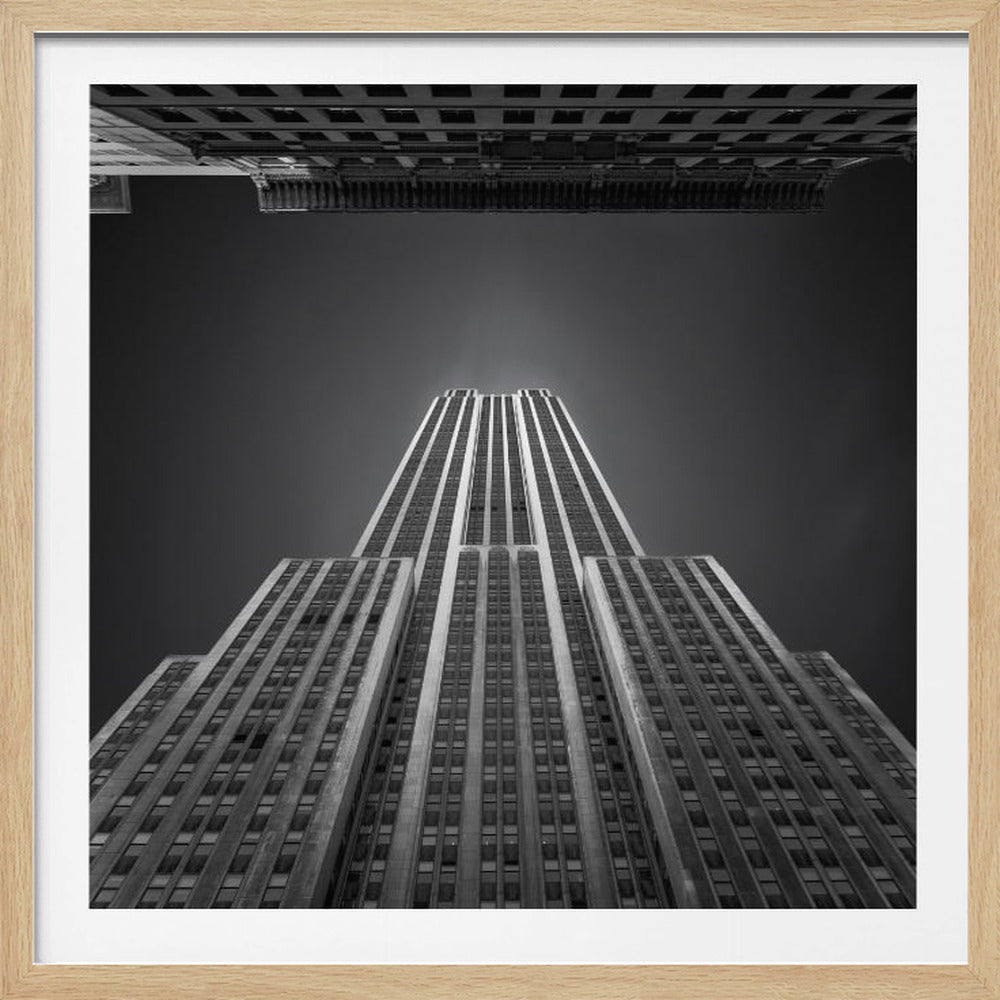 A dramatic, low-angle, black and white photograph of the Empire State Building, looking up from the street. The building's Art Deco lines stretch towards a dark grey sky, with another building visible at the very top of the frame, all enclosed in a light brown wood frame. Wall Art
