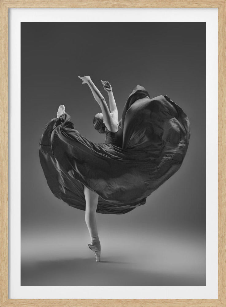 A dramatic black and white photograph of a ballerina captured mid-twirl. She is perfectly balanced on the tip of one pointe shoe, with her other leg kicked high. Her arms are raised elegantly above her head, and her long, dark dress billows out around her in a magnificent, fluid circle against a solid gray background. Wall Art