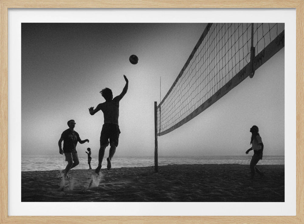 A black and white action shot of people playing volleyball on a beach, presented in a silver frame. The scene is silhouetted against a light sky, likely during sunrise or sunset. One player is captured mid-jump, arm raised to hit the ball over the net. Other players are visible on the sand, with the ocean in the background. Decor