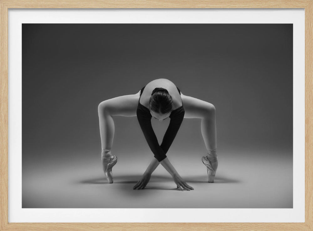 A framed black and white photograph of a ballerina in a dramatic and flexible pose. She is bent forward with her legs wide apart on pointe, her arms crossed in front of her with hands on the floor, creating a striking symmetrical shape against a plain gray background. Decor
