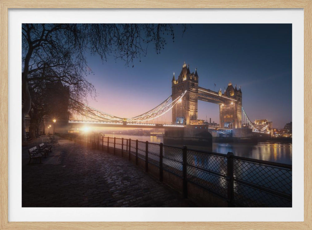 A scenic photograph of London's Tower Bridge at sunrise, its lights glowing warmly against a deep blue and orange sky. In the foreground, a cobblestone path follows the River Thames, leading the eye towards the iconic bridge and the rising sun. Poster