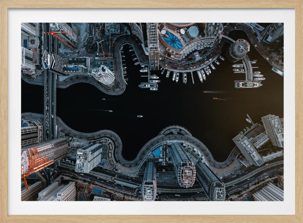A dramatic top-down aerial photograph of a sprawling modern city at night. A dark waterway curves through the center, surrounded by illuminated skyscrapers, busy roads, and a marina packed with boats, all within a silver picture frame. Artwork