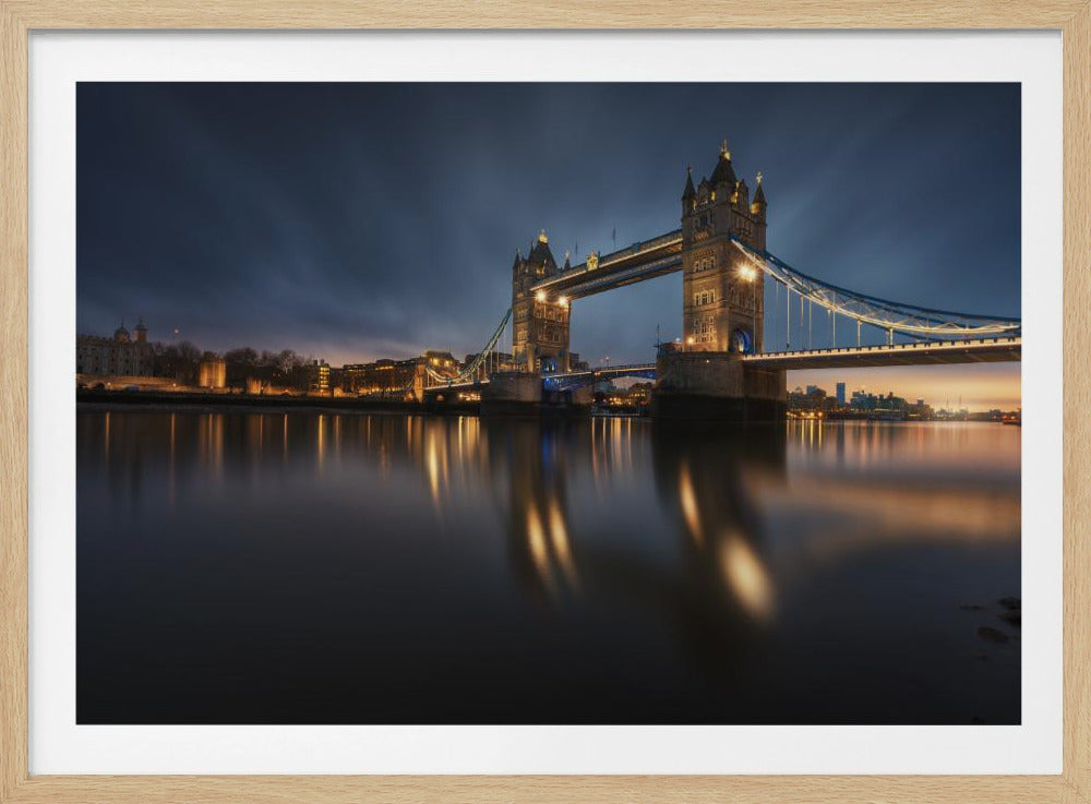 A long-exposure photograph of London's Tower Bridge at dusk, brilliantly illuminated with golden lights that cast shimmering reflections on the calm River Thames. The sky is a deep, dark blue, and the city skyline glows in the background, all enclosed within a silver frame. Print