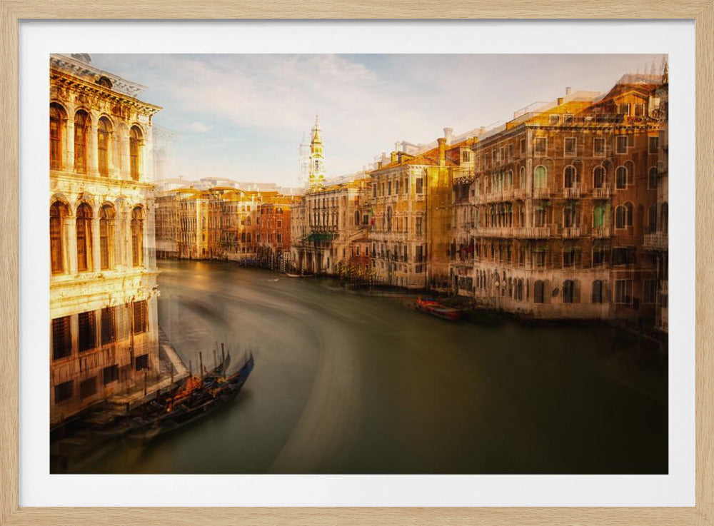 A long-exposure, artistic photograph of the Grand Canal in Venice, presented in a silver frame. The water is a smooth, dark blur, with motion streaks from gondolas. Ornate, historic buildings in shades of gold and brown line the canal under a pale blue sky, their reflections shimmering. The image has a dreamy, ethereal quality with soft motion blur throughout. Decor