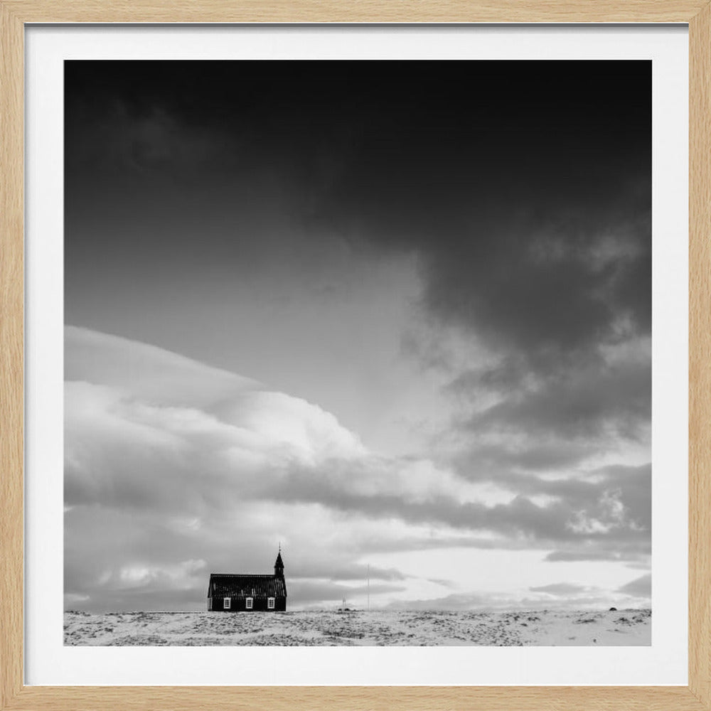 A framed black and white photograph of a small, solitary dark church on a snow-covered hill under a vast, dramatic sky with dark, moody clouds. Artwork