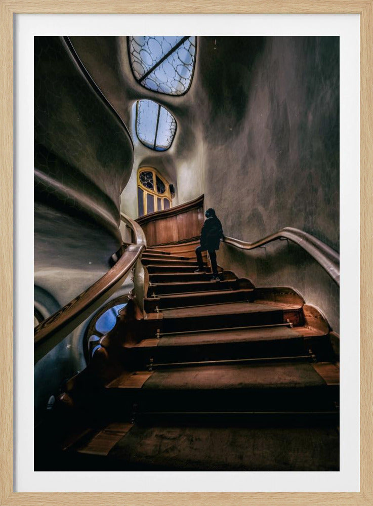 A low-angle view up a dark, winding wooden staircase inside a building with surreal, organic architecture. A person in a dark coat is walking up the stairs towards uniquely shaped windows that show a blue sky, contrasting with the dimly lit, textured gray walls. Print