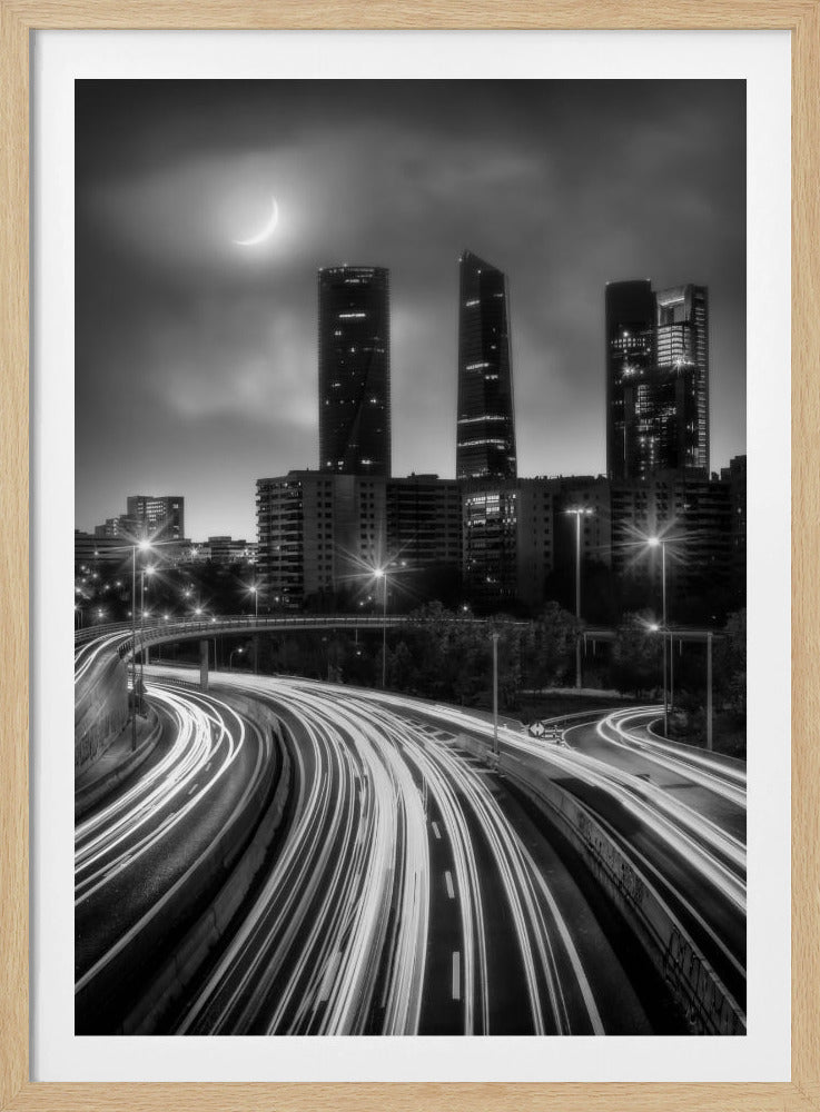 A dramatic black and white long-exposure photograph of a bustling city at night. Curving light trails from traffic fill the foreground, leading towards a modern skyline of illuminated skyscrapers under a cloudy sky with a crescent moon. Decor
