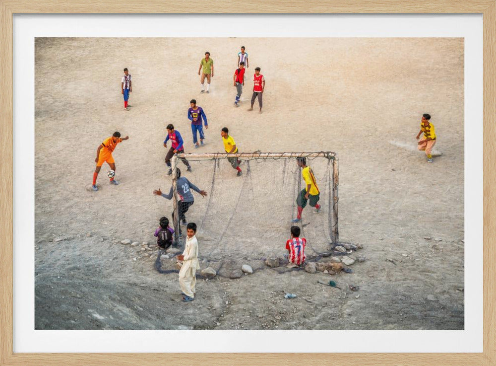 An overhead view of a group of boys and young men playing a spirited game of soccer on a large, dusty dirt field. A player in a bright orange uniform prepares to kick the ball towards a makeshift goal, defended by a goalie in a dark blue jersey. Other players in various colored shirts are scattered across the field, while a few young spectators watch from the sidelines. The entire image is enclosed in a silver frame. Wall Art