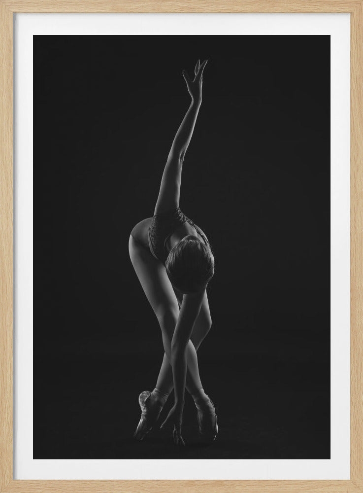 A dramatic black and white photograph of a ballerina posed against a dark background. She is bent forward at the waist, with her legs crossed and on pointe. One arm reaches up towards the ceiling in a graceful curve while the other hand touches the floor near her feet, showcasing her flexibility and strength. Wall Art