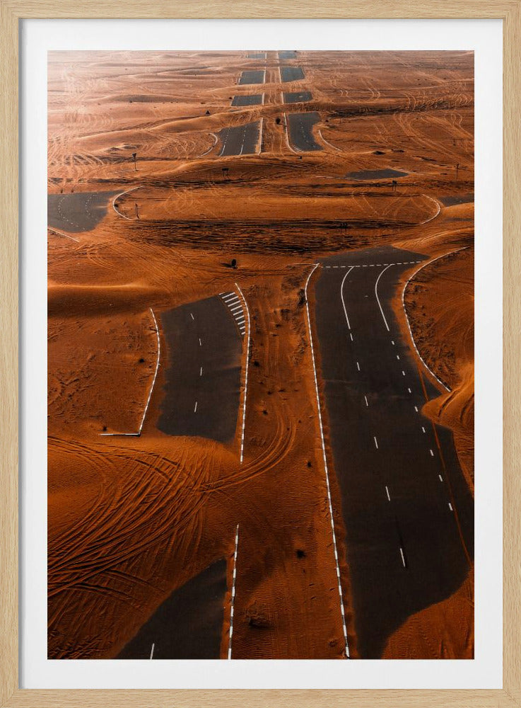 An aerial view of a multi-lane, black asphalt road being consumed by vast, rolling sand dunes of a deep orange color. The sand covers sections of the road, creating a surreal pattern of man-made lines and natural, windswept textures. Print