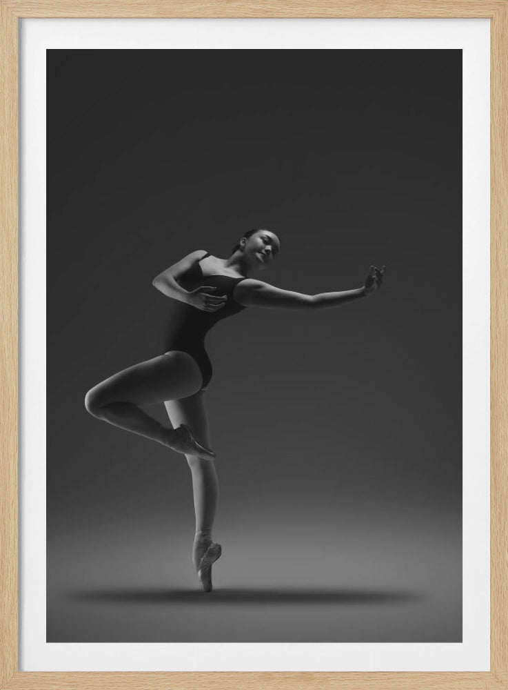 A dramatic low-angle, black and white studio photograph of a ballerina in a dark leotard. She is gracefully posed en pointe on one leg, with her other leg bent and raised. Her arms are extended elegantly, and the lighting highlights the contours of her body against a plain, dark gray background. The entire image is enclosed in a simple black frame. Artwork