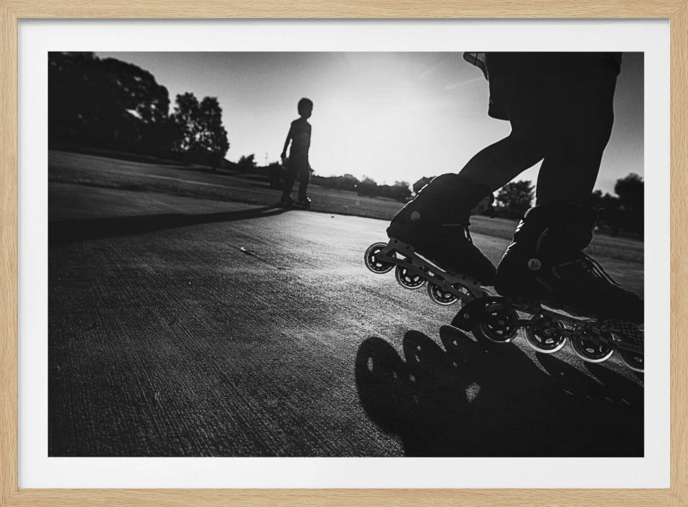 A low-angle, black and white photograph of two people rollerblading on a paved path at sunset. The foreground is a close-up of one person's rollerblades and their long shadow, while the second person is silhouetted in the distance against the bright sun. Artwork