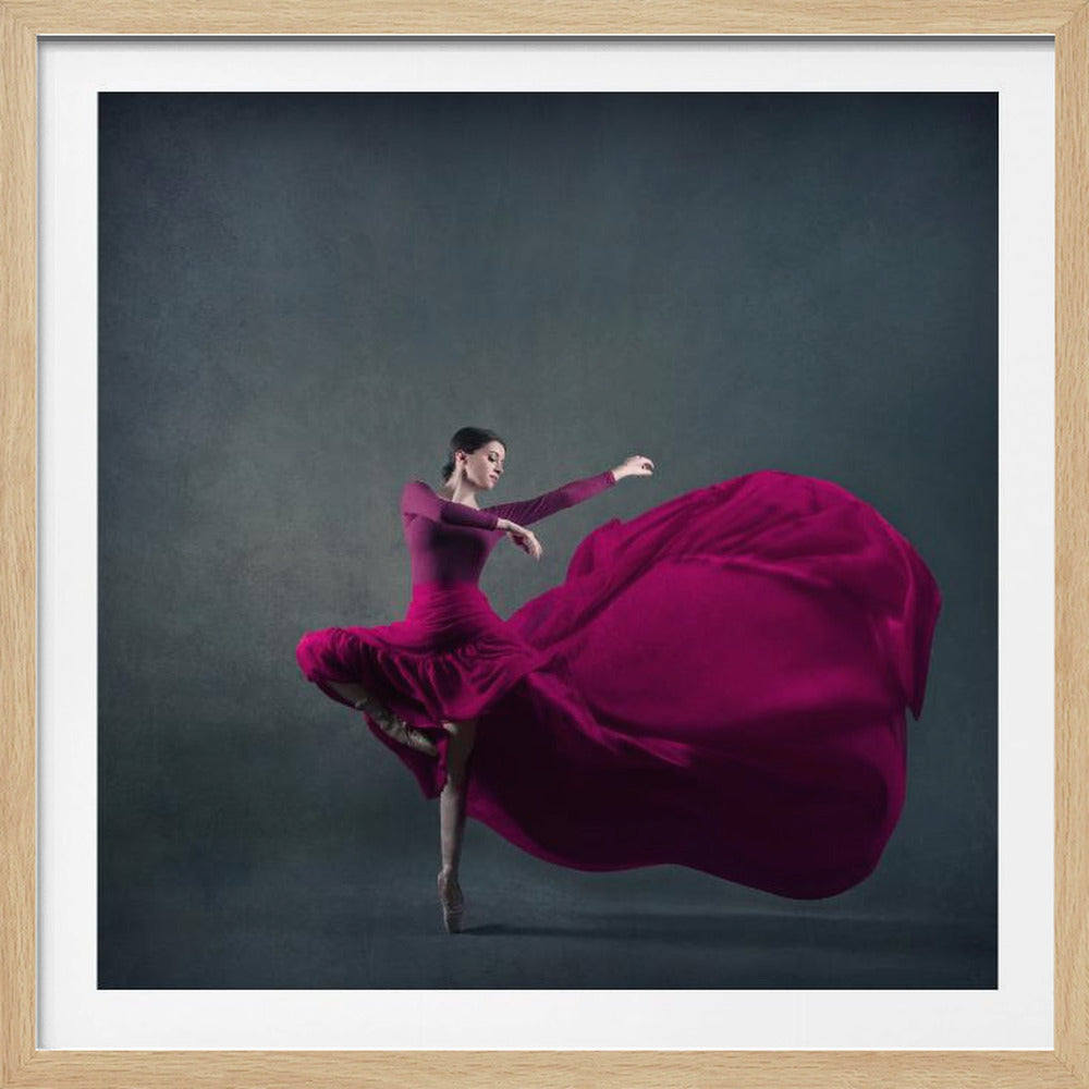 A graceful ballet dancer in a vibrant magenta dress strikes a pose on pointe, with the long, flowing fabric of her skirt billowing out dramatically behind her against a dark, textured gray background, all enclosed in a simple light wood frame. Decor