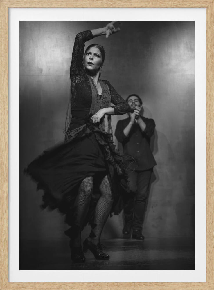 A dramatic black and white photograph of a female flamenco dancer in mid-performance, her skirt swirling as she moves. With one arm raised, her expression is intense and focused. In the soft-focused background, a male accompanist claps his hands. Decor
