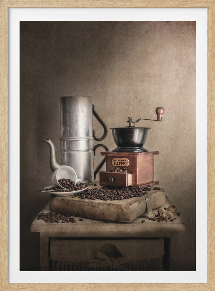 A vintage still life photograph featuring a classic aluminum Neapolitan coffee pot, a wooden hand-crank coffee grinder, and an overturned white cup spilling roasted coffee beans onto a rustic wooden surface against a textured brown background. Artwork