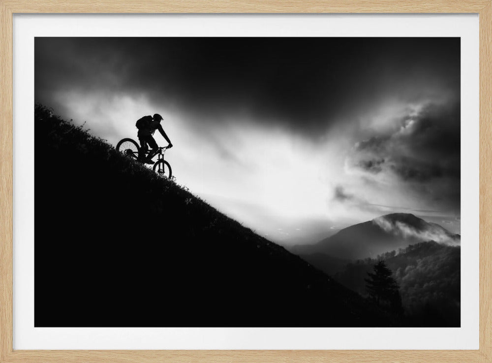 A striking black and white photo of a mountain biker's silhouette on a steep decline, set against a dramatic, light-filled cloudy sky with distant, misty mountains. Artwork