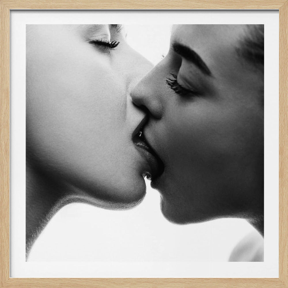 A close-up, black and white photograph of two women engaged in an intimate kiss. Their faces are shown in profile, with their eyes closed, against a bright white background. The image is enclosed in a light-colored wooden frame. Artwork