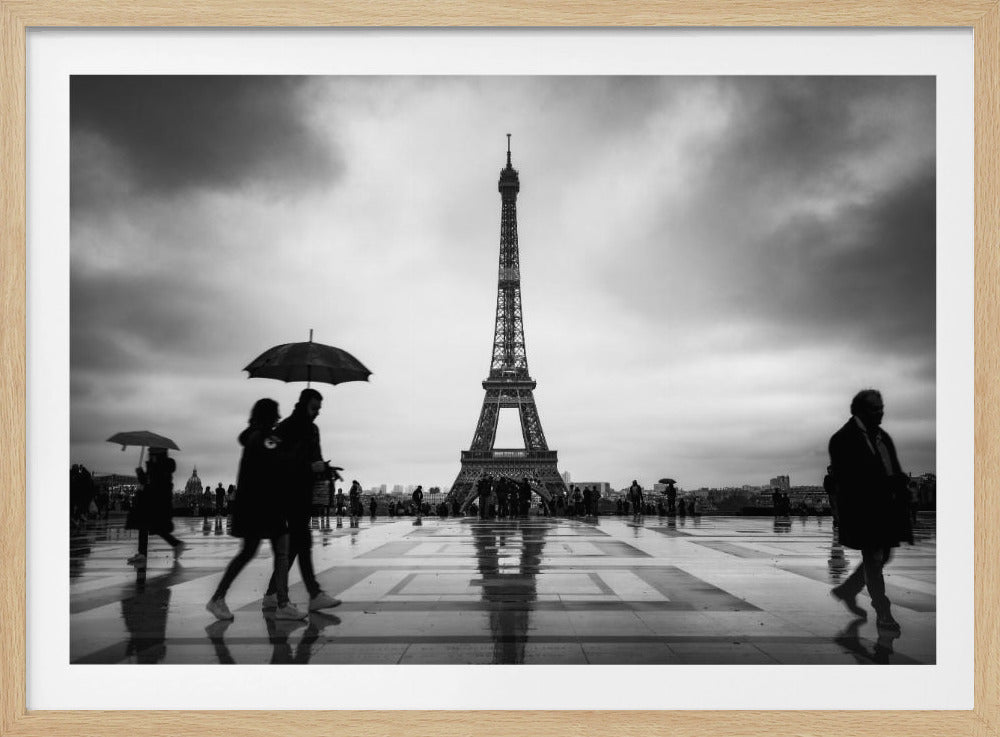 A black and white photograph of the Eiffel Tower on a rainy day. In the foreground, blurred figures of people walk across a wet, reflective plaza, some carrying umbrellas. The sky is overcast and dramatic. Print