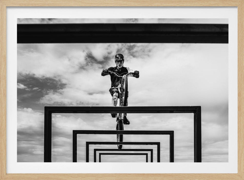 A striking black and white photograph taken from a low angle, showing a cyclist in full gear balancing on a mountain bike. The cyclist is positioned centrally above a series of black rectangular frames that create a receding, tunnel-like visual effect against a cloudy sky. The entire image is presented within a silver picture frame. Artwork