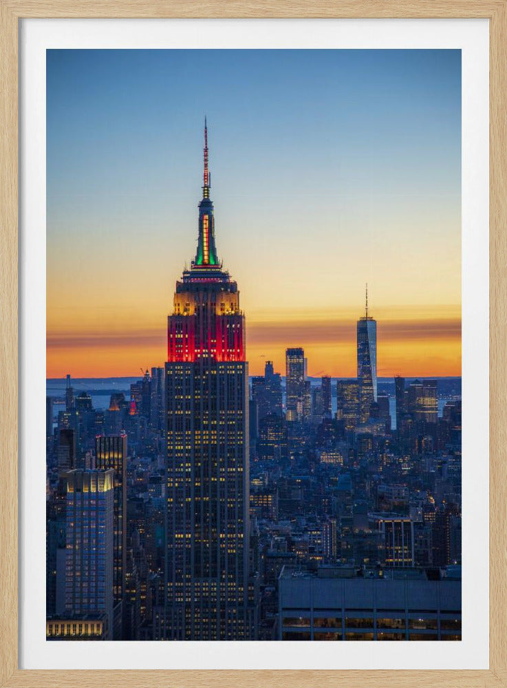 A vertical photograph of the New York City skyline at sunset, with the Empire State Building in the foreground. The building is illuminated with festive red and green lights on its upper floors, set against a sky that transitions from deep blue to a warm orange glow at the horizon. The surrounding cityscape, including the distant One World Trade Center, is bathed in the cool light of dusk, with scattered warm lights from building windows. Decor