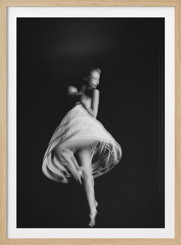 A black and white photograph capturing a dancer in motion against a dark background. The dancer, wearing a light-colored, translucent dress, is captured with a slow shutter speed, creating an ethereal blur that emphasizes the grace and movement of the twirling figure. Decor