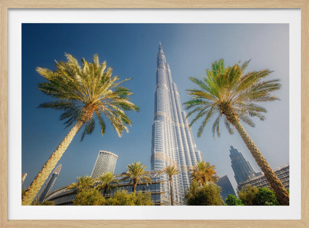 A low-angle photograph looking up at the towering Burj Khalifa skyscraper in Dubai, framed by two large palm trees against a clear, vibrant blue sky. Wall Art