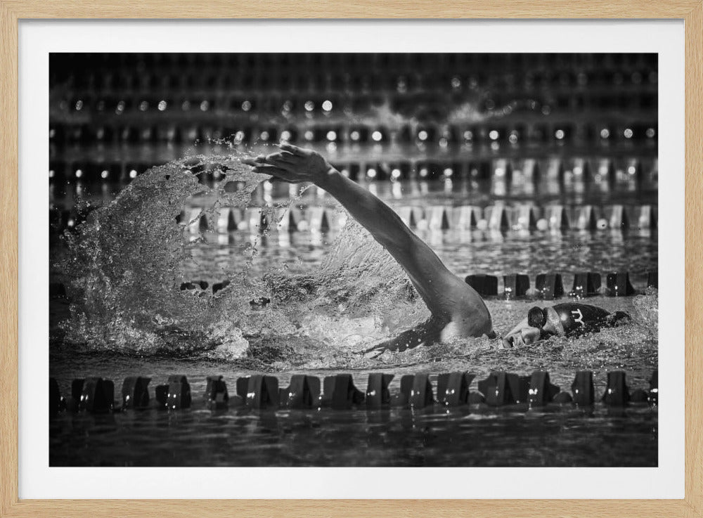A dramatic black and white action photograph of a competitive swimmer mid-stroke, captured within a silver frame. The swimmer's arm is extended forward, creating a powerful splash of water. The focus is on the motion and intensity of the sport, with the pool's lane lines blurred in the background. Wall Art