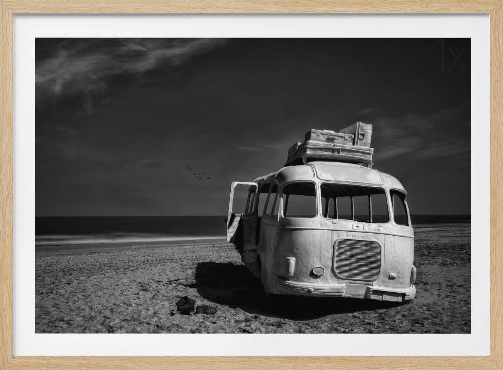 A dramatic black and white photograph of a vintage bus with luggage stacked on the roof, parked on a sandy beach. The ocean stretches out to the horizon under a dark, moody sky, with a small flock of birds flying in the distance. Print