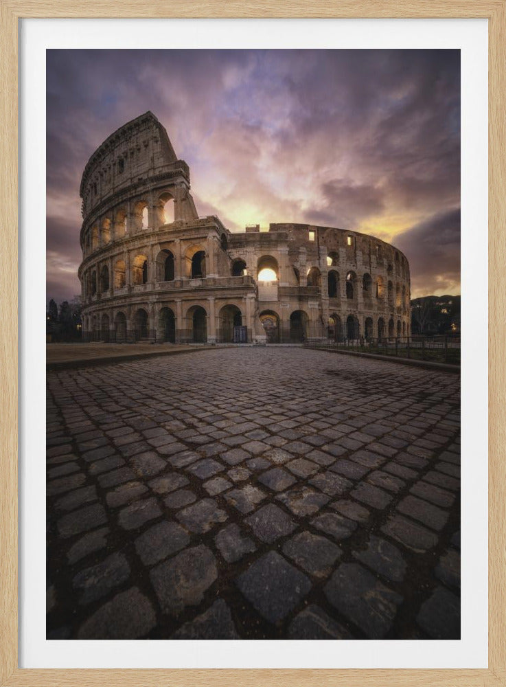 A dramatic low-angle photograph of the Colosseum in Rome at sunset. The foreground is filled with a dark cobblestone road leading towards the ancient amphitheater, which is bathed in the warm light of the setting sun against a moody, purple and orange cloudy sky. Decor