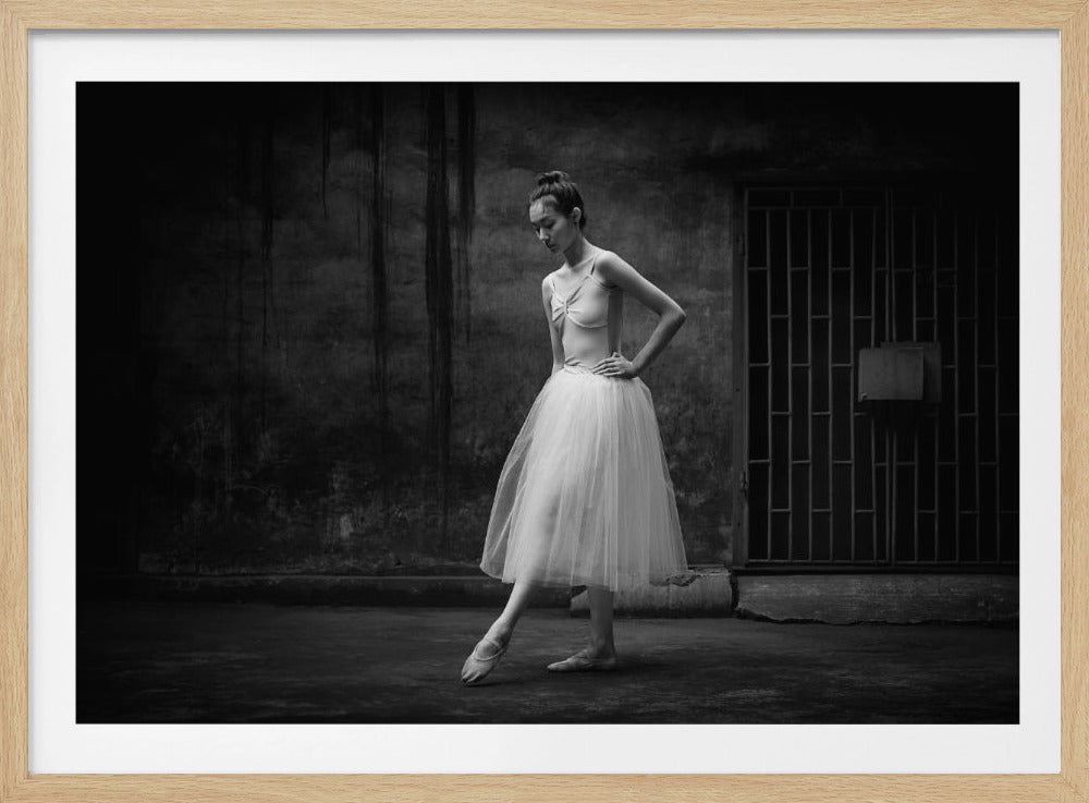 A framed black and white photograph of a ballerina in a white leotard and tutu. She stands in a graceful pose in a gritty, dark urban setting with a rustic wall and a barred gate behind her. Print