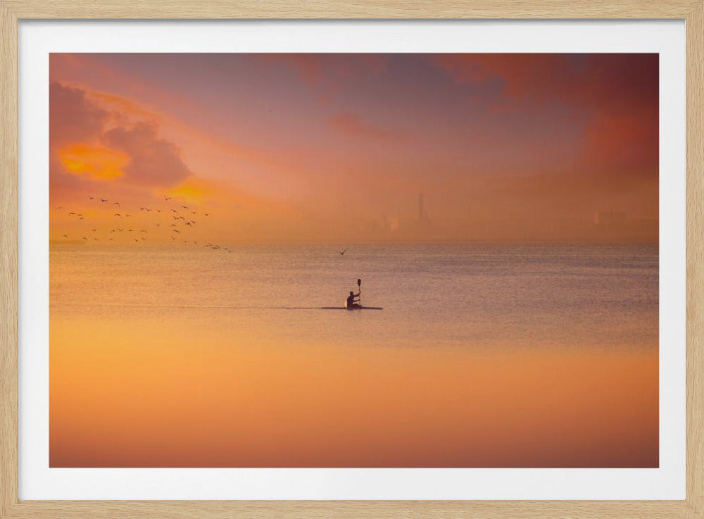 A serene photograph of a lone kayaker silhouetted against a vast expanse of calm water during a hazy, golden sunset. The sky and water are bathed in warm hues of orange, pink, and purple. A flock of birds flies in the distance, and the faint outline of a city skyline is visible on the horizon. Wall Art