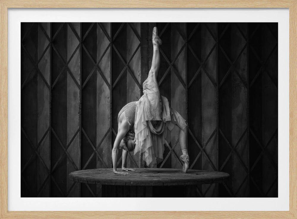 A framed, black and white photograph of a ballerina performing an extreme backbend on a round wooden table. One leg is extended straight up towards the ceiling while the other foot is on pointe on the table. She is wearing a leotard and a sheer skirt, and her hands are flat on the table. The background is a dark, geometrically patterned wall. Poster