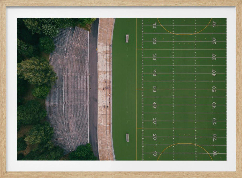 An aerial top-down photograph of an American football field beside empty, weathered concrete stadium bleachers. The vibrant green field is marked with white yard lines, and the gray stands are nestled amongst lush green trees. The entire scene is enclosed in a silver frame. Artwork