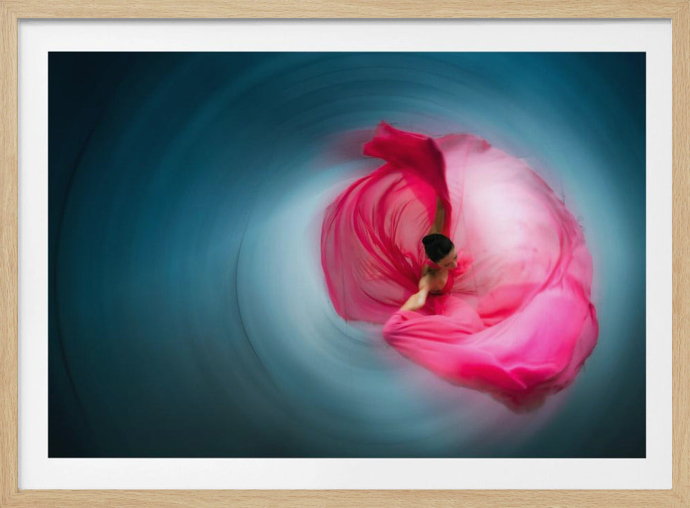 An overhead shot of a dancer in a vibrant, flowing pink dress, spinning against a radially blurred blue background. The motion creates a swirl of color, with the dancer at the heart of the vortex. The artwork is presented in a silver frame. Artwork