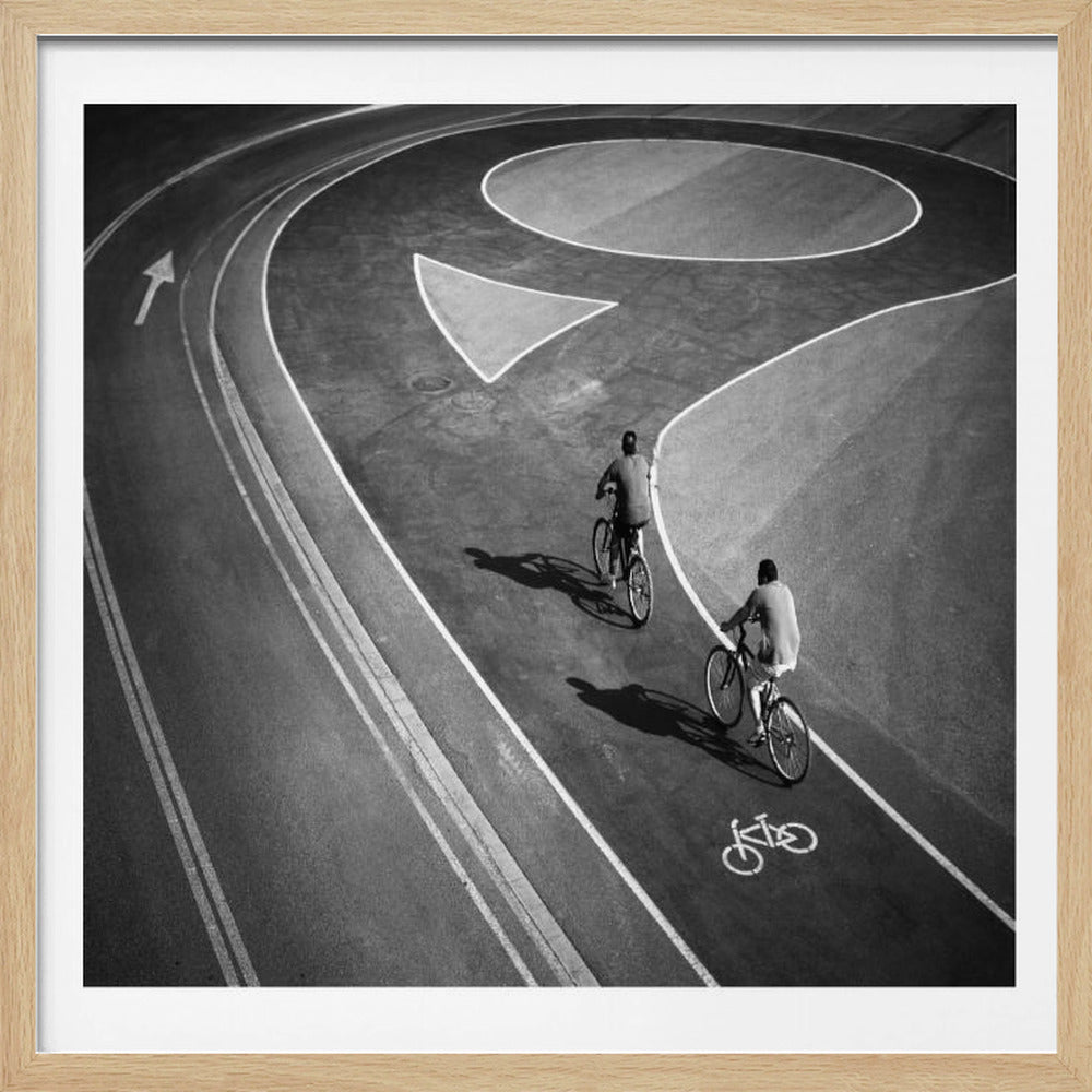 A framed, high-angle black and white photograph of two people cycling on a curving asphalt bike path. The path is marked with bold white lines, including a large arrow and a bicycle symbol, creating a strong graphic pattern. The cyclists cast long shadows on the pavement. Poster