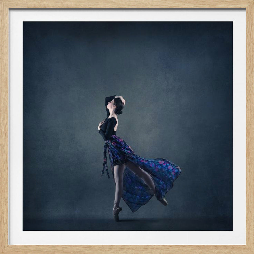 A full-length portrait of a graceful ballerina posing against a dark blue-grey background. She is on pointe, wearing a black top and a flowing blue and purple floral skirt that is caught in motion. Her hands are elegantly placed on her chest and head, expressing emotion. The image is presented in a light wood frame. Decor