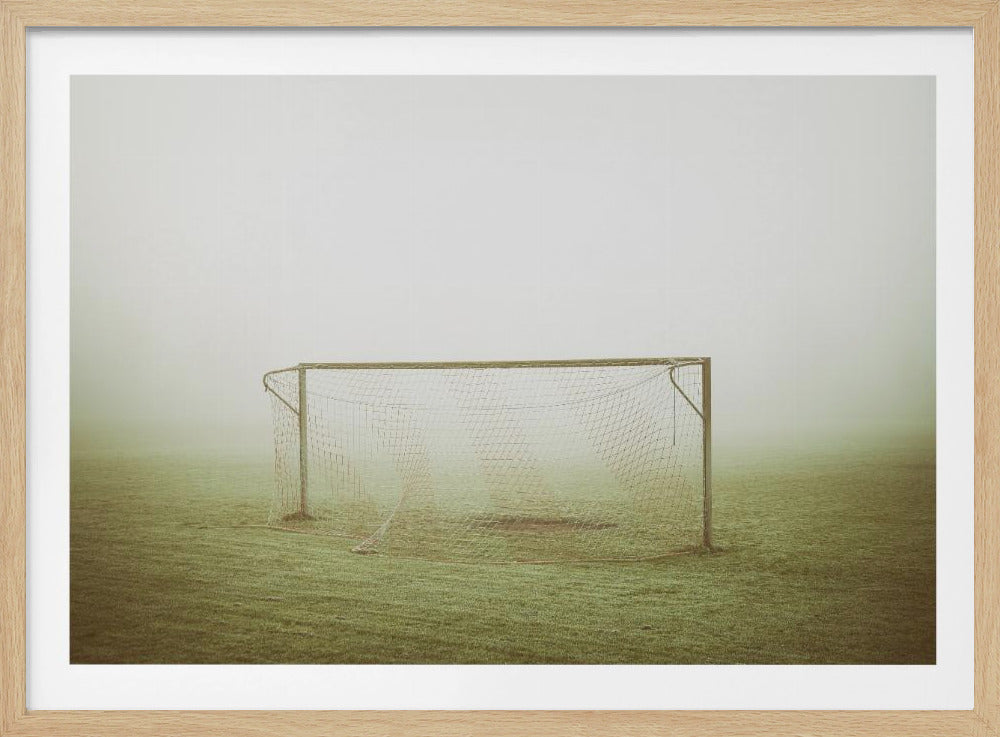 A framed photograph of an empty soccer goal with a net, standing alone on a green grass field enveloped in thick, white fog, creating a minimalist and atmospheric scene. Poster