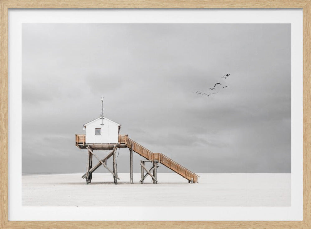 A minimalist, desaturated photograph of a small white cabin on tall wooden stilts on a vast, empty beach. A long wooden staircase leads up to the cabin. The sky is a flat, overcast grey, and a flock of dark birds is visible flying in the distance. Print