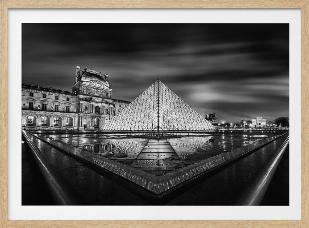 A dramatic black and white night photograph of the illuminated Louvre Pyramid in Paris, with its reflection shimmering on the wet ground. The historic Louvre Palace stands in the background under a moody, cloudy sky, and the entire scene is enclosed in a silver frame. Wall Art