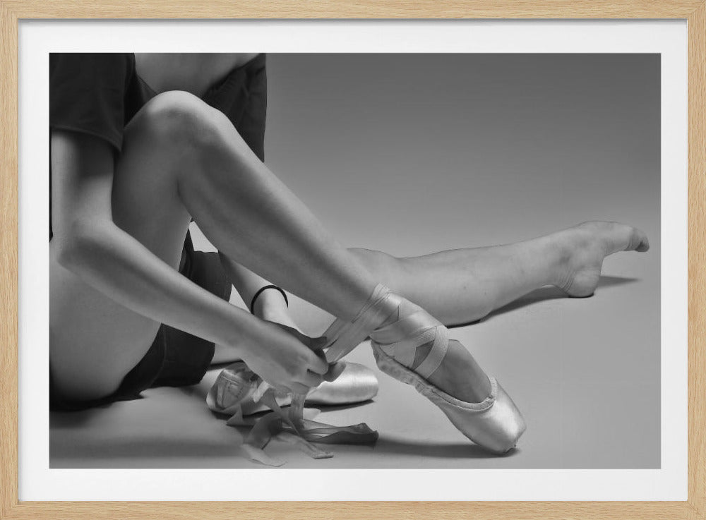 A black and white close-up photograph of a ballet dancer sitting on the floor, carefully tying the ribbons of a satin pointe shoe on her right foot. The image has a simple silver frame and captures a quiet moment of preparation. Poster