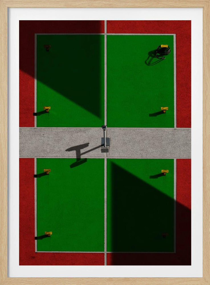 A high-angle, symmetrical photograph of a green and red sports court, viewed from directly above. Strong, sharp shadows cut diagonally across the composition, creating a geometric and abstract pattern of light and dark. A net post in the center casts a T-shaped shadow on the concrete dividing strip. Print