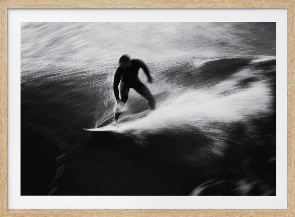 A dynamic black and white photograph of a surfer in a wetsuit carving through a wave. The image uses motion blur to emphasize the speed and energy of the water, creating a powerful and artistic effect. The artwork is presented in a simple silver frame. Print