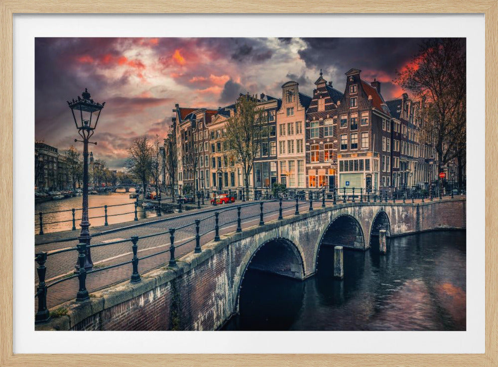 A dramatic sunset over a classic Amsterdam canal, featuring a brick arched bridge in the foreground, traditional canal houses lining the water, and a vintage lamppost on the left. Print