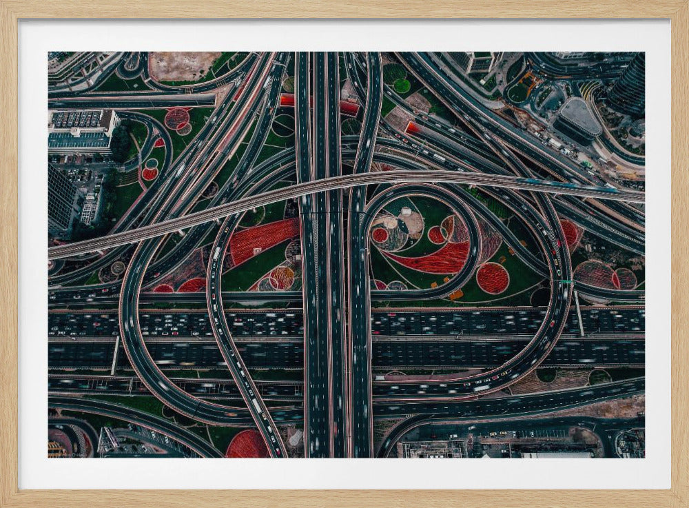 An aerial, top-down photograph of a sprawling and complex highway interchange, framed in silver. Multiple levels of roads and overpasses curve and intertwine, with blurred streaks of traffic visible. The spaces between the highways feature manicured green lawns and artistic red and patterned landscaping. Wall Art