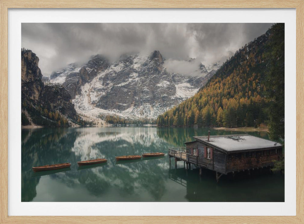 A framed photograph of a serene mountain lake scene. A rustic wooden boathouse sits on stilts over the calm, greenish water, with several empty rowboats floating nearby. In the background, majestic snow-dusted mountains are partially obscured by low-hanging clouds, and a hillside is covered in golden autumn trees, all reflected on the lake's surface. Wall Art