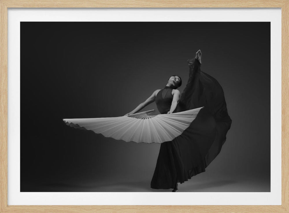 A framed, black and white photograph of a female dancer in a dramatic pose. She wears a dark, flowing gown and holds a large, open white fan in front of her. One leg is kicked high in the air as she arches her back, creating a sense of graceful movement against a simple dark gray background. Print