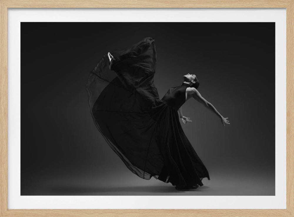 A dramatic black and white photograph of a female dancer in a flowing black gown, captured mid-motion. She is arched backwards with her head tilted up, her arms extended gracefully. The fabric of her dress billows up and around her, creating a sense of dynamic movement against a solid dark gray background. The image is presented within a silver frame. Decor