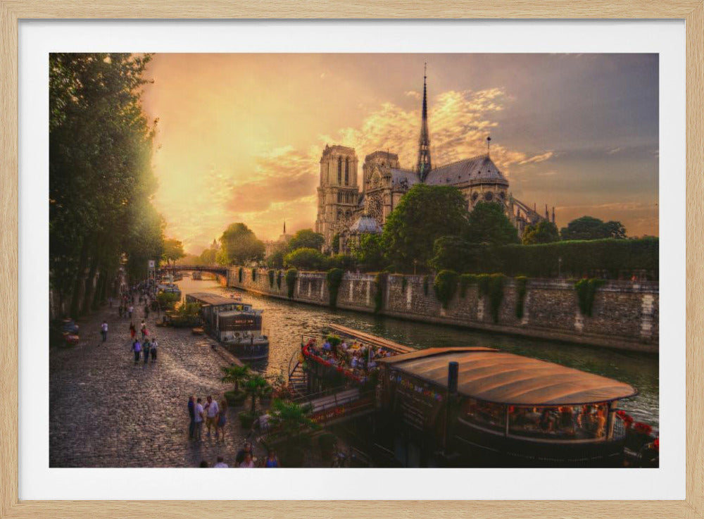 A scenic photograph of the Notre Dame Cathedral in Paris during a warm, golden sunset, as viewed from across the River Seine. People are strolling along the cobblestone riverbank and riding on tour boats docked on the water. The image is presented within a silver picture frame. Decor